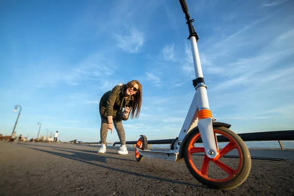 Chica atractiva y su scooter en el asfalto. Chica joven sonriendo y mostrando símbolo de victoria. Patinete scooter blanco con ruedas naranjas. Actividad deportiva. Momentos felices — Foto de Stock