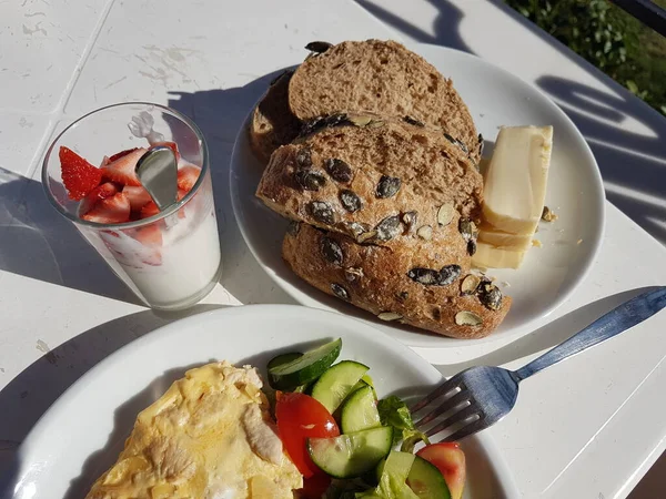 Desayuno saludable en el balcón. Pan integral con semillas de calabaza y queso. Ensalada de pepinos frescos y tomates. Higos frescos. Fresas con yogur. Por encima de la luz solar directa — Foto de Stock