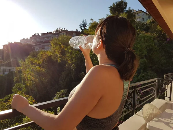 Молодая спортсменка пьет свежую воду из бутылки на солнечном балконе, освежаясь — стоковое фото