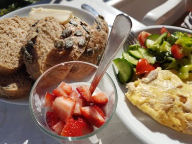 Balkonda sağlıklı bir kahvaltı. Kabak çekirdeği ve peynirli tam tahıllı ekmek. Salatalık ve domatesli salata. Taze incir. Yoğurtlu çilek. Doğrudan güneş ışığının üzerinde