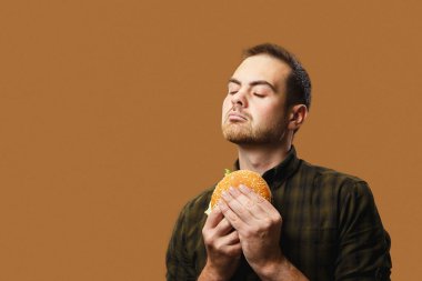 Genç bir adam büyük bir burgeri iştahla tutar. Turuncu arkaplan, kopyalama alanı