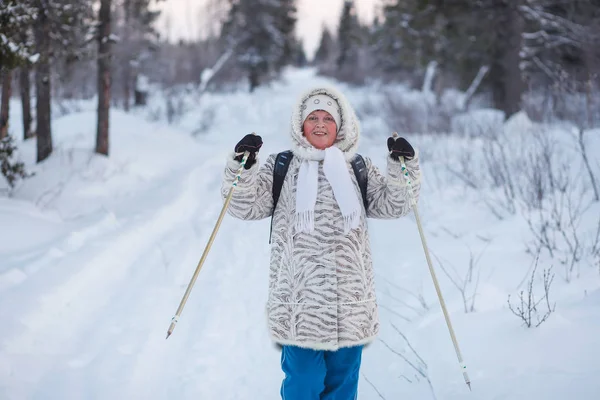 Mutlu, güzel, yaşlı bir kadın kış ormanında İskandinav yürüyüşü yapıyor. Şenlikli bir yaşam tarzı, temiz hava ve iyi bir ruh hali. — Stok fotoğraf