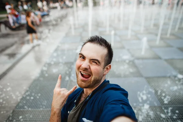 Un hombre feliz y atractivo se toma una selfie en una fuente seca en el parque. Un hombre exitoso muestra un gesto de victoria —  Fotos de Stock