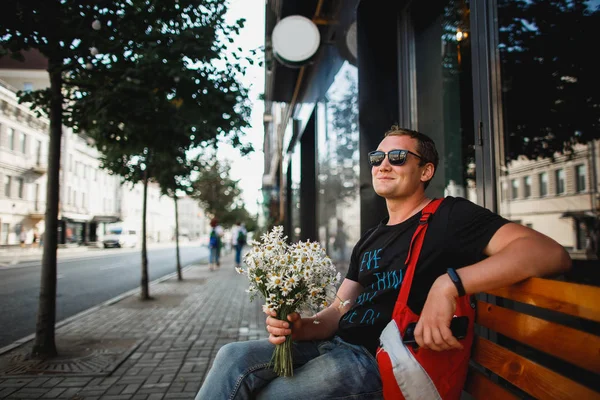 Hombre alegre se sienta en un banco con un ramo de margaritas esperando una cita —  Fotos de Stock