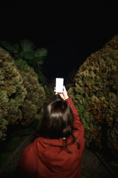 A nő telefonnal éjjel a parkban hátulról néz. Sötét sikátor van előttünk. — Stock Fotó
