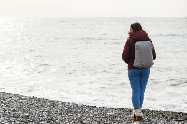 Una mujer en una chaqueta con una mochila observa las olas del mar por detrás — Foto de Stock