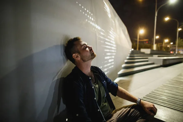 Человек сидит и думает на современной ночной набережной в свете фонарей — стоковое фото