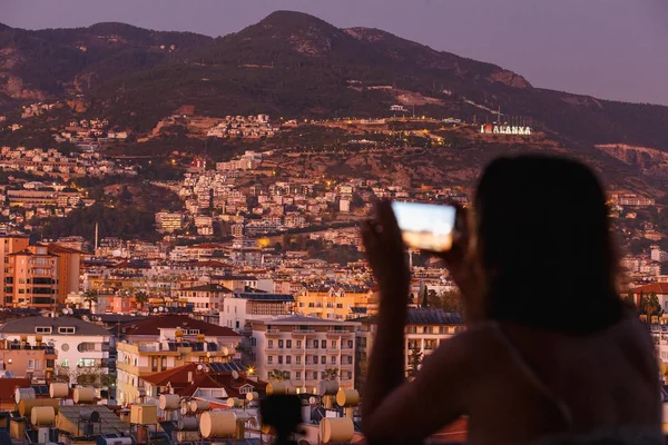 Female traveller make a panoramic photo of the mountain cityscape from the balcony to her phone for social networks. Travel to Alanya, Turkey. Summer vacation. Dusk time. Focus on the background — Stock Photo, Image