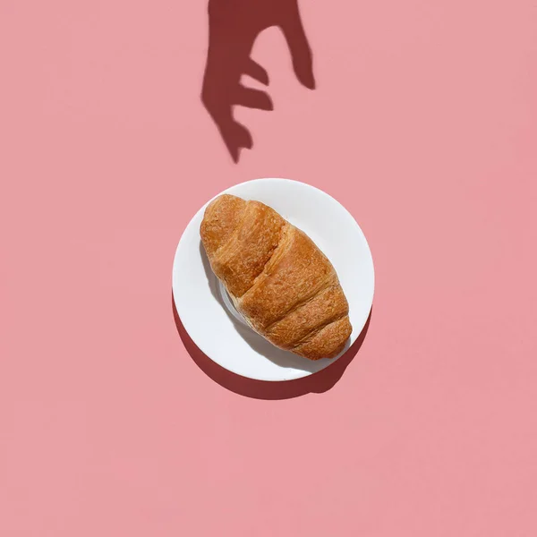 A sombra da mão de mulheres consegue um grande croissant em um contexto rosa. Diretamente acima do plano leigos — Fotografia de Stock