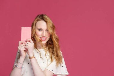 Neşeli sarışın kız elinde pembe bir defter tutuyor, kameraya bakıyor ve gülümsüyor, pembe bir arka planda soyutlanmış.