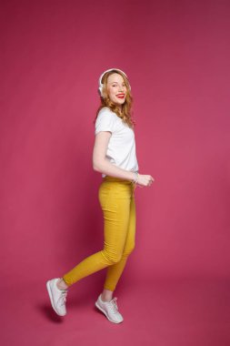 Beyaz tişörtlü, sarı pantolonlu ve beyaz kulaklıklı güzel sarışın kadın pembe arka planda müzik dinliyor. Dinamik stüdyo fotoshoot