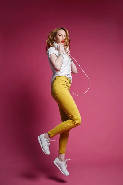 白いTシャツ、黄色のズボンと白いヘッドフォンの美しいブロンドの女性はピンクの背景に音楽を聴く。ダイナミックスタジオフォトシュート — ストック写真