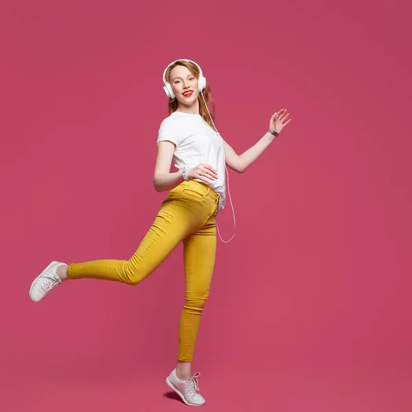 Mulher loira bonita em camiseta branca, calças amarelas e fones de ouvido brancos escuta música em fundo rosa. Fotografia de estúdio dinâmica — Fotografia de Stock