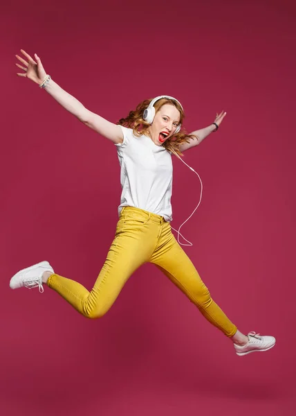Krásná blondýna v bílém tričku, žlutých kalhotách a bílých sluchátkách poslouchá hudbu na růžovém pozadí. Dynamické studiové focení — Stock fotografie