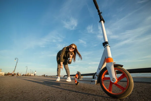 Chica atractiva y su scooter en el asfalto. Chica joven sonriendo y mostrando gesto de victoria. Patinete scooter blanco con ruedas naranjas. Verdaderas emociones — Foto de Stock