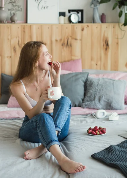 Mujer sexy rubia con una taza de té o café y fresas divertirse en la cama. Llevaba sujetador blanco y vaqueros casuales. Buenos días. — Foto de Stock