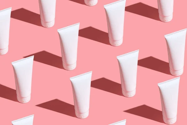 Açık pembe masada beyaz krema tüpleri. Yüzüne, ellerine, bacaklarına ve derisine dikkat et. Kadın güzellik ürünleri. Kozmetik desen. Şişelerde logo için boş bir yer. Dümdüz uzanan sert ışık