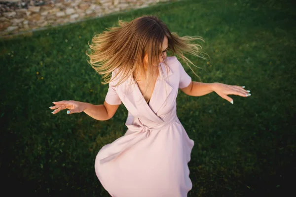 Menina alegre bonita cai em uma dança na grama verde. Um vestido rosa delicado flutters lindamente. Retrato dinâmico de cima — Fotografia de Stock