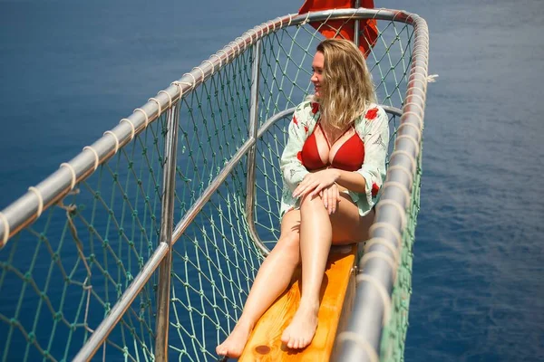 Curvy woman in a red swimsuit sitting on the front of the boat deck on turqouise blue sea background. View from the back - Sea voyage or cruise concept — Stock Photo, Image