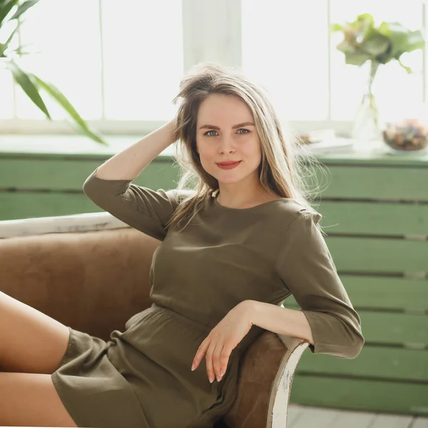 Schöne Frau in einem khakifarbenen Kleid im grünen Interieur. Charismatische Frau amüsiert sich im kurzen Kleid auf dem Sofa. Schöner Tag zu Hause. — Stockfoto