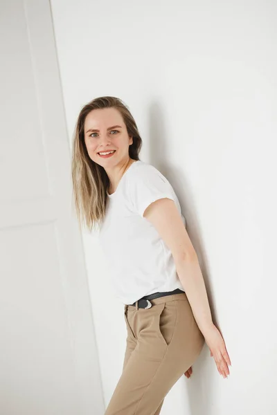 Retrato emocional de una hermosa mujer caucásica activa en el interior de la pared blanca, mirando a la cámara. Ella se vistió con camiseta blanca y pantalones beige — Foto de Stock