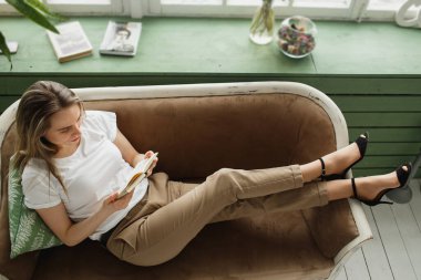 Güzel kadın pencerenin yanındaki kanepede uzanmış kitap okuyor.