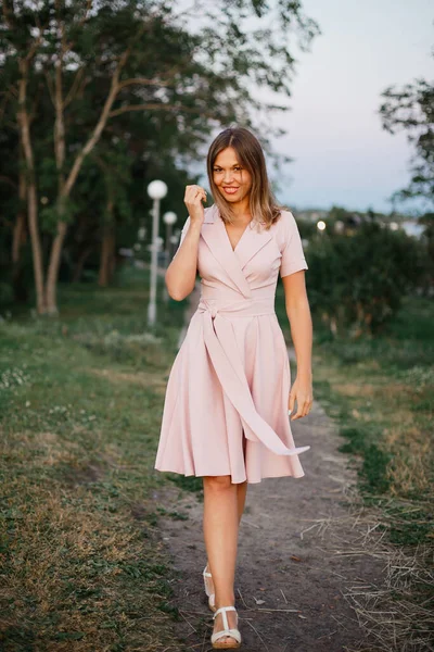Игривая молодая женщина в розовом летнем платье наслаждается прогулкой в весеннем парке вечером — стоковое фото