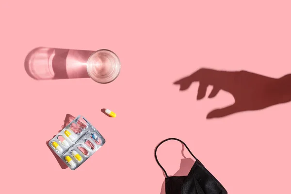 Pills in a blister, a glass of water, black medical mask and a hand shadow on a pink background. Flat lay, top view — Stock Photo, Image