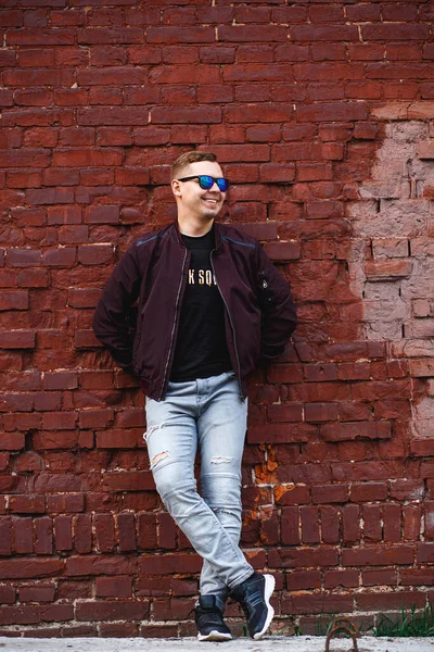 Молодий чоловік в джинсах, кросівки і піджак стоїть своїми руками в кишенях біля цегляної стіни — стокове фото
