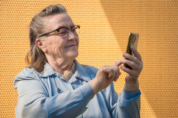 Портрет старушки, бабушка делает селфи на открытом воздухе в солнечный день. Современный бабушкин палец коснулся сенсорного экрана ее телефона. У неё смартфон в руке. стиль жизни — стоковое фото