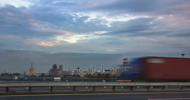 Zeitraffer Stadtbild bei Sonnenuntergang mit der Bewegung von Wolken und verschiedenen Gebäuden im Hintergrund und dem Autoverkehr auf einer Autobahn im Vordergrund — Stockvideo