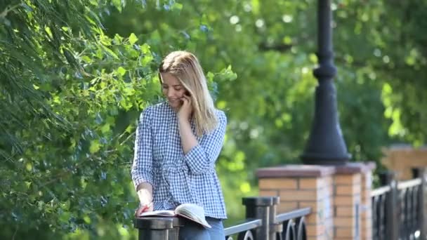 Ung flicka i trädgården pratar i telefonen och att göra anteckningar i hennes anteckningsbok. — Stockvideo