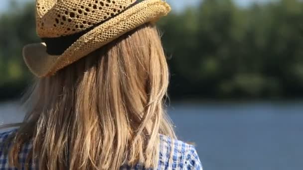 Primer plano de una chica en un sombrero de paja mira al río — Vídeos de Stock