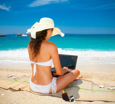 Dizüstü bilgisayarlı, şapkalı güzel bir kadın sahilde eğleniyor. Yunanistan.