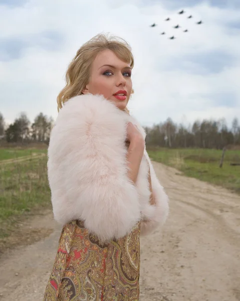 Genç sarışın kadın bej elbise ve kırsal yolda kısa pembe kürk manto