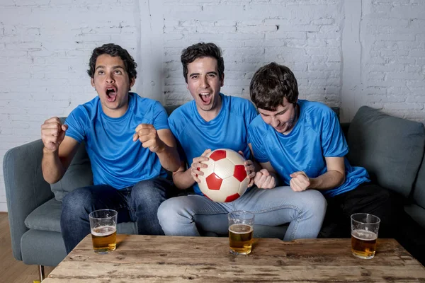 Excited group of young football fans sitting on sofa watching football game and drinking beer