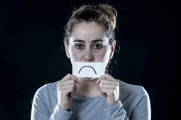 Young beautiful sad and depressed latin woman covering her sad face with a paper cut out sadder face in a depression concept on isolated black background.