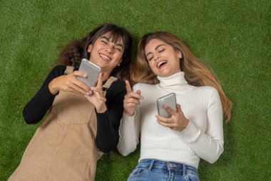 Mutlu kız arkadaşlar sosyal medya uygulamasında cep telefonu sohbeti yapıyor ya da internetten alışveriş yapıyor. İki kadın, çim parkında uzanmış akıllı telefonlarda birlikte eğleniyor. İnsanların yaşam tarzı ve teknolojisi.