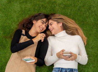 Mutlu kız arkadaşlar sosyal medya uygulamasında cep telefonu sohbeti yapıyor ya da internetten alışveriş yapıyor. İki kadın, çim parkında uzanmış akıllı telefonlarda birlikte eğleniyor. İnsanların yaşam tarzı ve teknolojisi.
