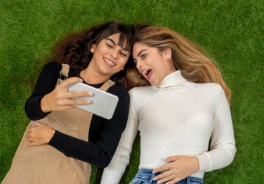 Mutlu kız arkadaşlar sosyal medya uygulamasında cep telefonu sohbeti yapıyor ya da internetten alışveriş yapıyor. İki kadın, çim parkında uzanmış akıllı telefonlarda birlikte eğleniyor. İnsanların yaşam tarzı ve teknolojisi.