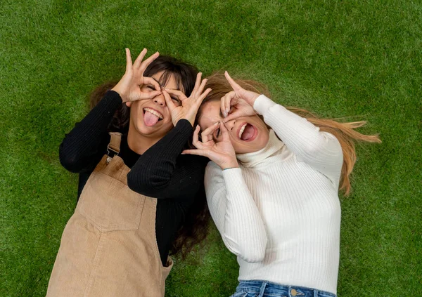 Top view of two stylish girl friends having fun laughing and making faces lying down on grass. Blonde and brunette happy young teenagers women enjoying time together. Friendship, love and care.