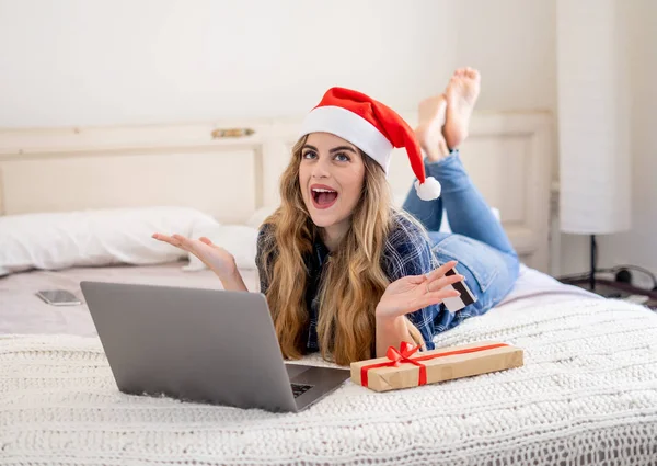 在网上购物和寒假期间 戴着圣诞礼帽的女人拿着信用卡 在家里用笔记本电脑选购圣诞礼物 兴奋地看着网上销售和信用卡设施 — 图库照片