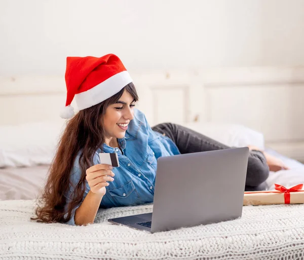 在网上购物和寒假期间 戴着圣诞礼帽的女人拿着信用卡 在家里用笔记本电脑选购圣诞礼物 兴奋地看着网上销售和信用卡设施 — 图库照片