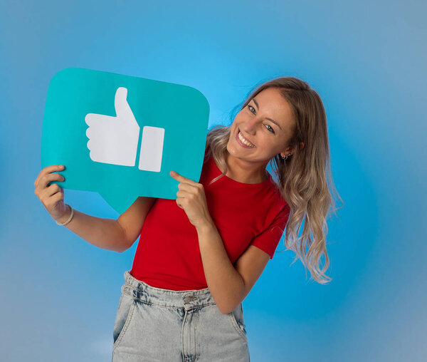 Beautiful young woman holding Like symbol social media notification icon asking followers to like her online comments, blog or business. In Internet obsession, networking and technology connections.