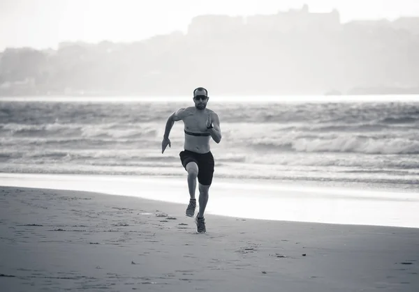 Athlete runner running with heart rate monitor and smart watch on beach. Shirtless fitness man exercising in outdoor training. Advertising style in sports technology healthcare wellness innovation.