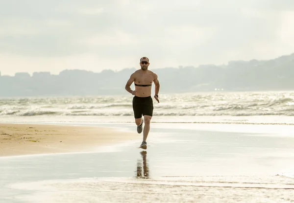 Athlete runner running with heart rate monitor and smart watch on beach. Shirtless fitness man exercising in outdoor training. Advertising style in sports technology healthcare wellness innovation.