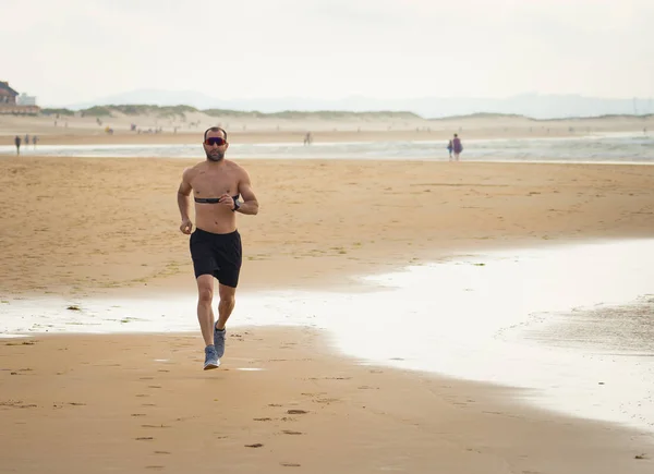 Sports fitness man with heart rate monitor running on beach with smart watch. Fit body male runner doing jogging workout at the sea in sport exercise and technology and healthy lifestyle concept.