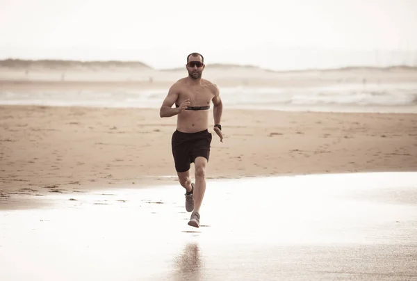 Sports fitness man with heart rate monitor running on beach with smart watch. Fit body male runner jogging by the seashore in workout exercise and training technology innovation in healthy lifestyle.