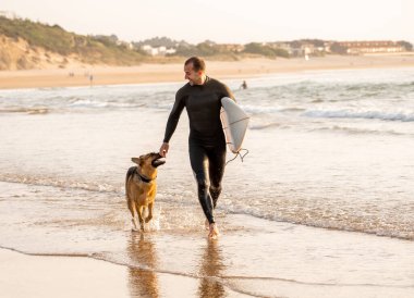 Sörfçü en iyi arkadaşı Alman çoban çalışan ve gün batımında köpek dostu plajda oynarken eğlenmek. Köpek, evcil hayvan dostu gezi ve açık macera yaşam tarzı ile yaz eğlenceli sörf tatil.