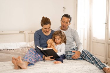 Baba, anne ve küçük kızı birlikte kitap okuyorlar. Mutlu bir ailenin evde masal okuyarak eğlendiği bir hayat tarzı. Mutlu aile, ebeveynlik ve eğitim konseptinde.